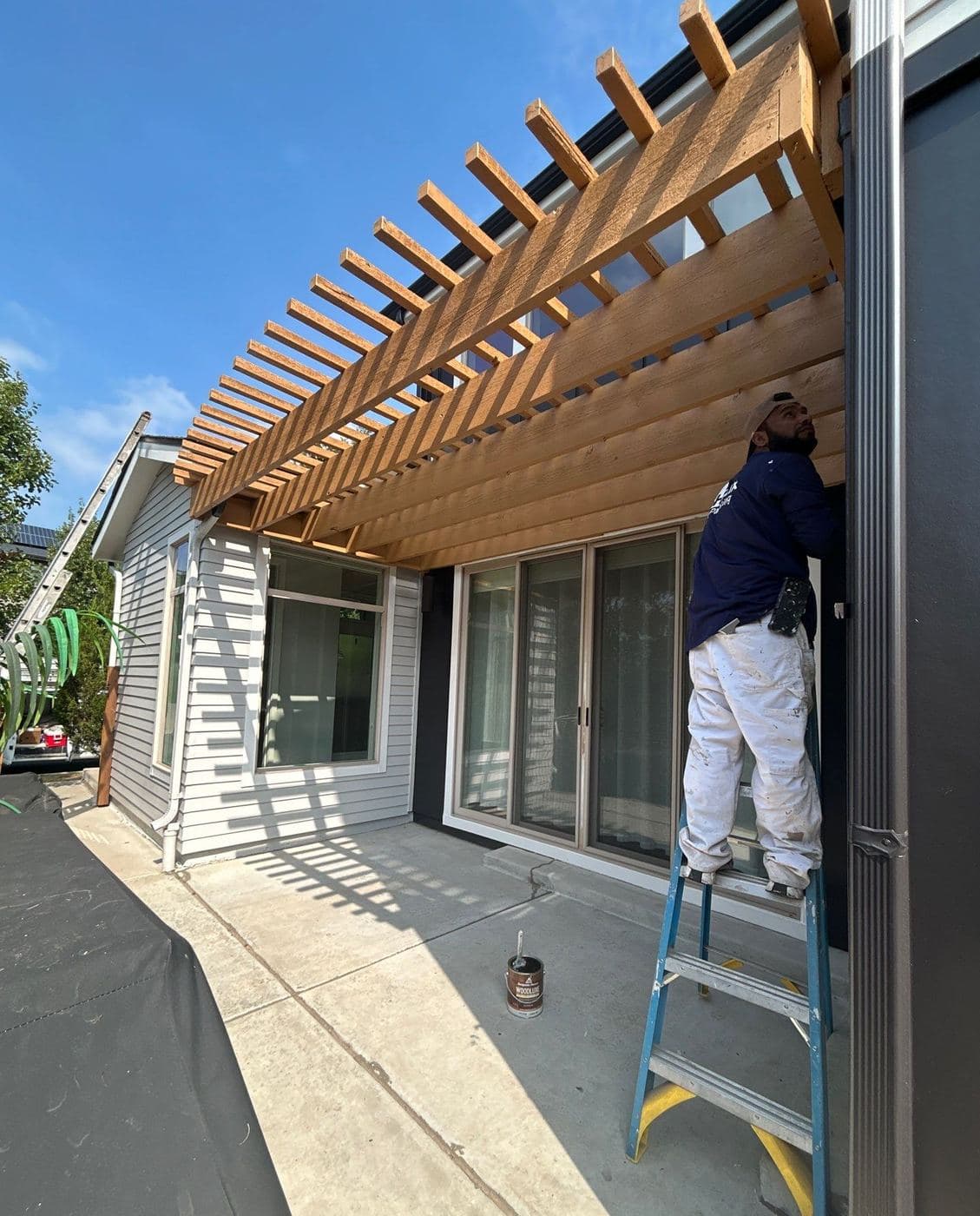 Painter on a ladder finishing a pergola beside a patio